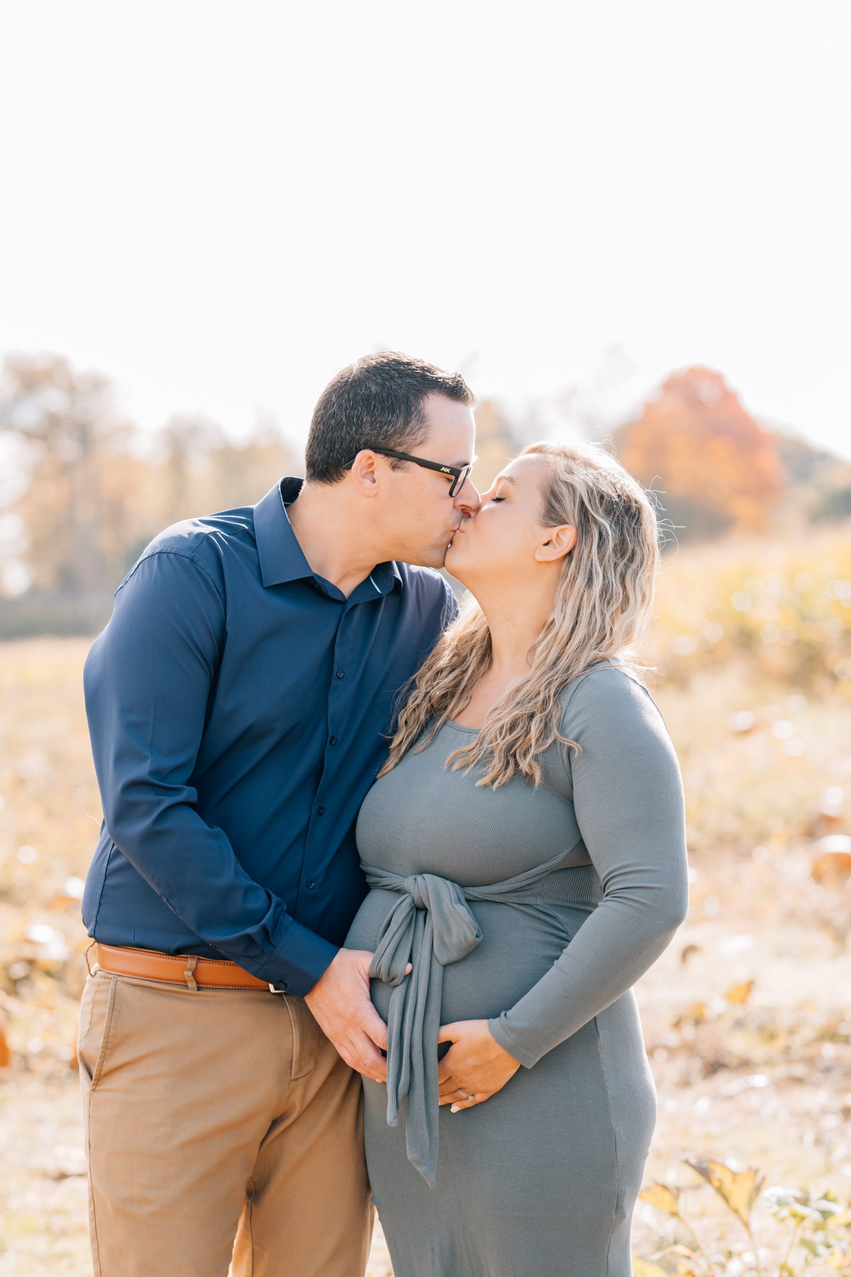Flemington Fall Mini Session | Maternity Photoshoot NJ | The D'Angiolillo Family mom and dad kiss in pumpkin patch holding hands on baby bump