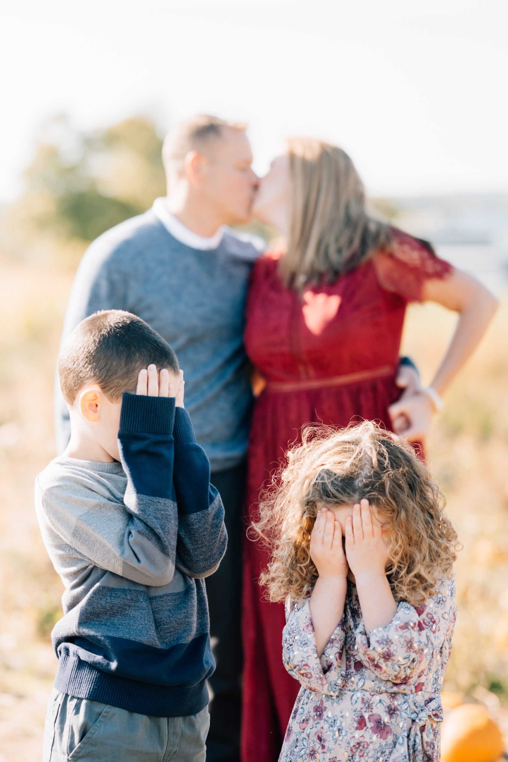 Family of 4 mom and dad kissing with kids covering eyes during fall mini portrait session at Pumpkin Junction at Everitt Farms in Flemington, NJ Ringoes, NJ with Motherhood and Family Photographer Anne Haug of Golden Heart Photography.