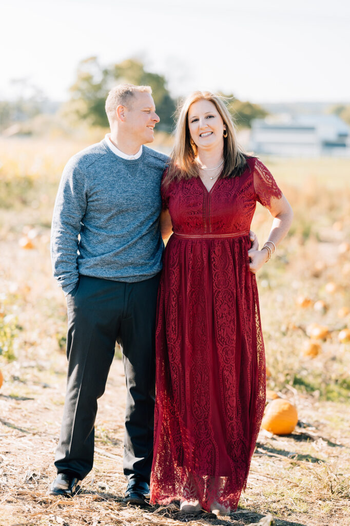 mom and dad pose together in pumpkin patch dad looking at mom in beautiful crimson dress during fall mini portrait session at Pumpkin Junction at Everitt Farms in Flemington, NJ Ringoes, NJ with Motherhood and Family Photographer Anne Haug of Golden Heart Photography.