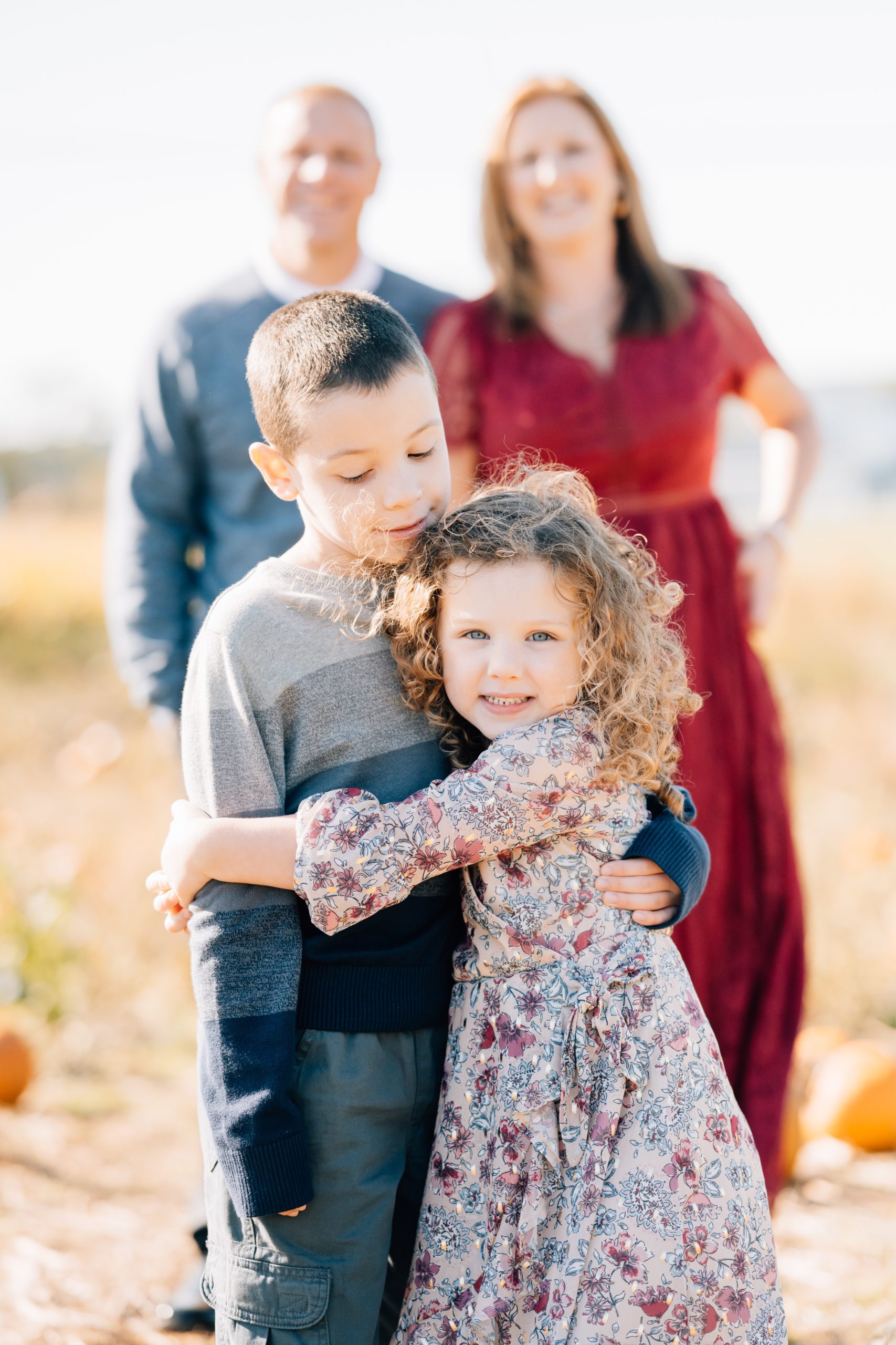 Family of 4 walks through pumpkin patch brother and sister hugging with mom and dad in background during fall mini portrait session at Pumpkin Junction at Everitt Farms in Flemington, NJ Ringoes, NJ with Motherhood and Family Photographer Anne Haug of Golden Heart Photography.